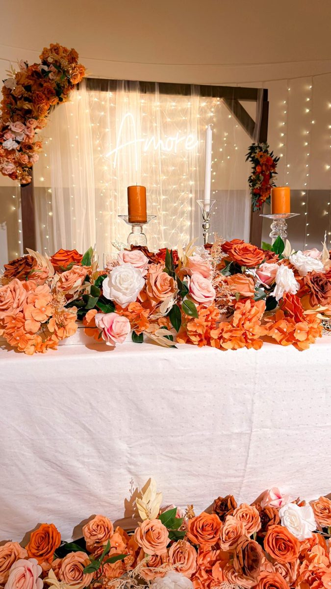 Amore-neonskilt bak lysgardin med blomsterbue og bord pyntet med oransje og rosa roser